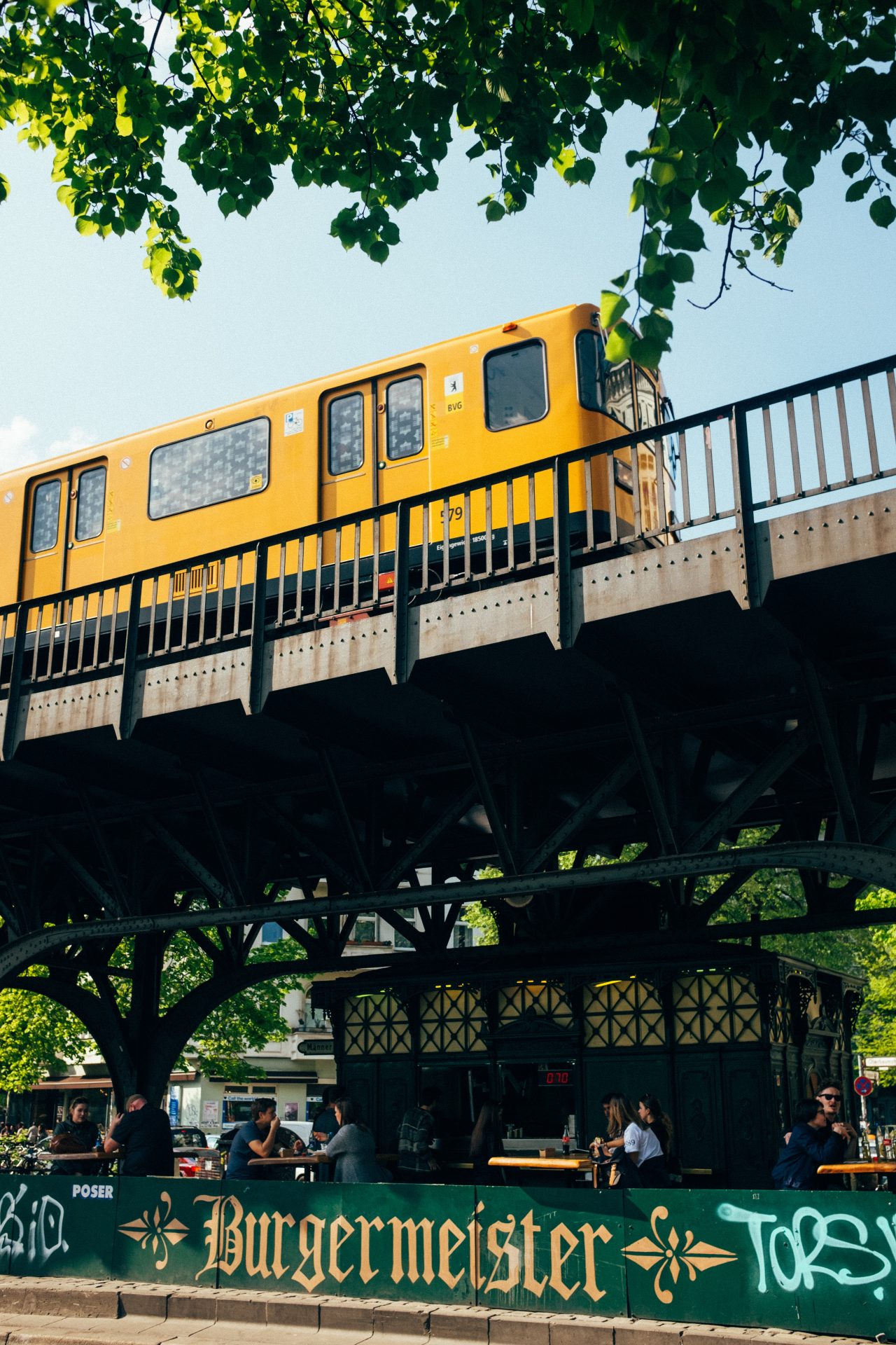Eine gelbe U-Bahn fährt über eine Brücke, unter der ein Häuschen ist und Menschen an Tischen essen. Auf einer Mauer steht 
