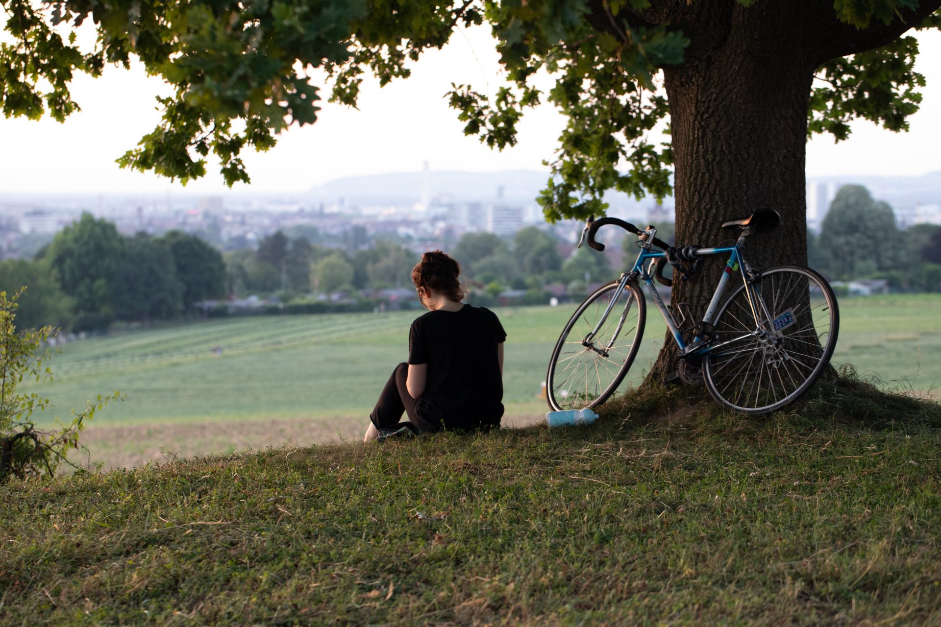 Eine Person sitzt unter einem Baum auf einer grünen Wiese. Am Baum lehnt ein Fahrrad.